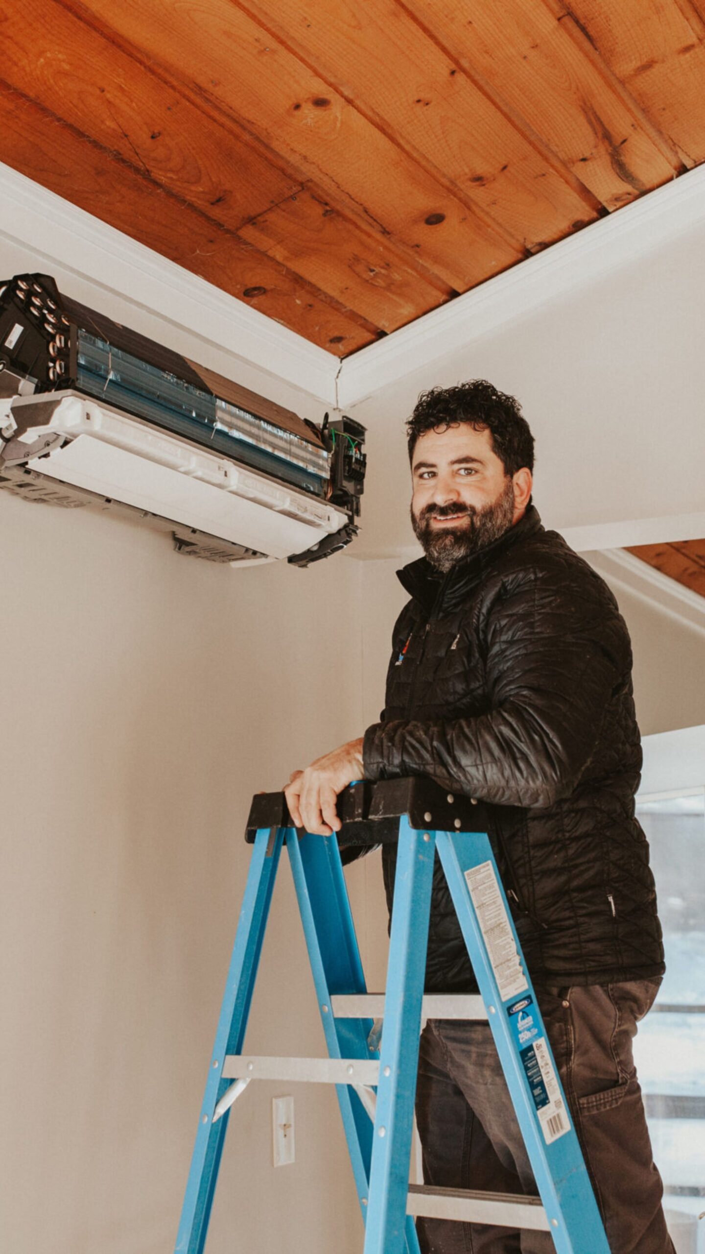 Ryan on ladder HVAC technician standing on a blue ladder installing a ductless mini split system in a home with wood-paneled ceiling. He is smiling and wearing a black insulated jacket.